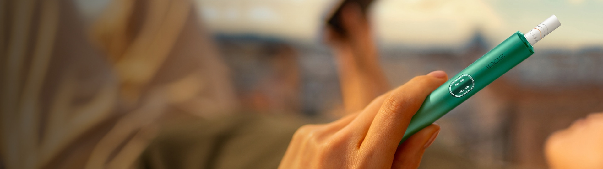 A woman holds an IQOS device while looking at a laptop.